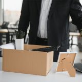 Employee packing up desk items into a cardboard box during a downsizing and layoffs process in the workplace.